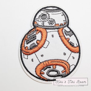 May include: Embroidered patch featuring the Star Wars droid BB-8. The patch has a white background with orange and black details. The design includes the droid's head and body, with a black border. The patch is from Kiki's Tiki Room.