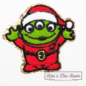 Pode incluir: Um alienígena verde de três olhos vestindo um traje de Papai Noel vermelho com um chapéu de Papai Noel branco e acabamento de glitter dourado. O alienígena está sorrindo e tem um cinto preto.