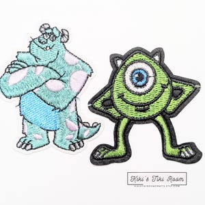 May include: Two embroidered iron-on patches featuring characters from the movie Monsters Inc. The patch on the left is blue and green and features Sully, a large, furry monster with one eye. The patch on the right is green and features Mike, a small, one-eyed monster with a wide grin.