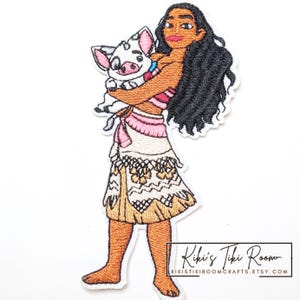 Peut inclure: Un écusson brodé représentant un personnage aux longs cheveux noirs tenant un porcelet blanc aux oreilles roses. Le personnage porte un haut beige et une jupe blanche et beige à motifs géométriques. Le texte en bas indique Kiki's Tiki Room.