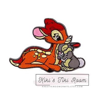 May include: Embroidered iron-on patch featuring Bambi the deer from Disney's Bambi, holding Thumper the rabbit. Bambi is orange and red with white spots, and Thumper is gray.