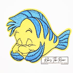 Op de afbeelding: Geborduurde patch met een gele en blauwe cartoonvis. De vis heeft blauw haar en vinnen, met een glimlachende uitdrukking. De patch heeft een zwarte rand en de tekst "Kiki's Tiki Room" is zichtbaar.