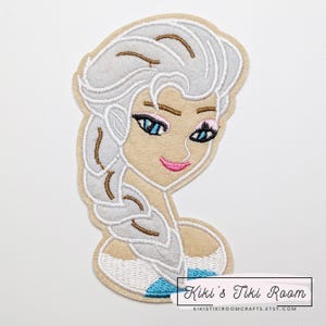 May include: Embroidered patch featuring a stylized image of a woman with long, silver hair in a braid. The patch has a tan background with blue eyes, pink lips, and the text "Kiki's Tiki Room" at the bottom.