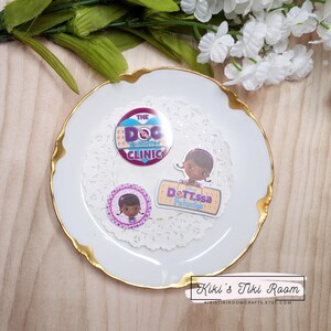 Puede incluir: Un plato blanco con un borde dorado, decorado con una servilleta de encaje blanca, presenta tres pegatinas redondas con personajes del programa de televisión infantil Doc McStuffins. Las pegatinas muestran a Doc McStuffins, Dottie y el logotipo de la clínica Doc McStuffins. El texto en las pegatinas dice "The Doc McStuffins Clinic" y "Dottie's Patchwork".
