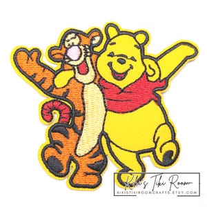 May include: Embroidered patch featuring Winnie the Pooh and Tigger. Winnie the Pooh is yellow with a red shirt, and Tigger is orange with black stripes. The patch has a yellow border and the text "Kiki's Tiki Room" at the bottom.