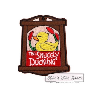 Op de afbeelding: Gestikt patch met een bruin houten frame en een geel eendje erin. De tekst "The Snuggly Duckling" staat in rode letters op een rode achtergrond.