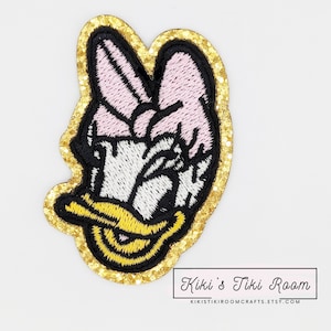 May include: A gold glitter iron-on patch featuring a cartoon duck with a pink bow. The duck has black eyes, a yellow beak, and a white body. The patch is approximately 2 inches in diameter.