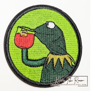 May include: A circular embroidered patch featuring a green frog character drinking from a red glass with a yellow accent. The design is set against a bright lime green background with a thick black border. The text Kiki's Tiki Room is visible.