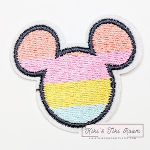 May include: An embroidered iron-on patch in the shape of Mickey Mouse's head. The patch is made of felt and features a rainbow color scheme with pink, yellow, and blue stripes. The patch is outlined in black and has a white background.