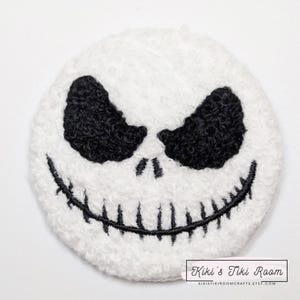 May include: White fuzzy fabric patch with a black embroidered Jack Skellington face. The face has two black eyes, a black nose, and a black stitched smile.
