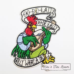 May include: Embroidered patch featuring a rooster playing a lute. The rooster has a red comb, yellow beak, and green feathers, wearing a blue shirt. The patch includes the text "Oo-De-Lally" and "Golly What a Day".