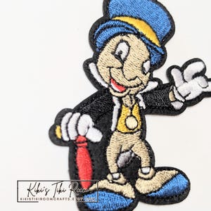 May include: An embroidered patch featuring a cartoon cricket character wearing a blue top hat with a yellow band, a black tailcoat, a yellow vest, and blue shoes. The character holds a red umbrella and has white gloves. Text reads Kiki's Tiki Room.