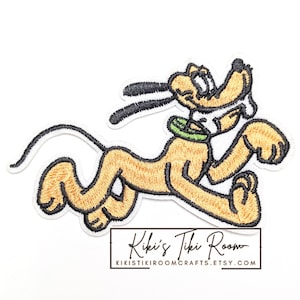 May include: Embroidered patch featuring a cartoon dog in a running pose. The dog is primarily yellow with black accents on the ears and nose. It has a green collar and a white outline. The patch includes the text "Kiki's Tiki Room" and a website address.