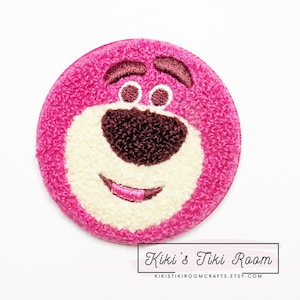 Peut inclure: Patch en peluche brodé rose et blanc d'un personnage de dessin animé avec un nez brun et une bouche souriante. Le personnage est du film Toy Story.