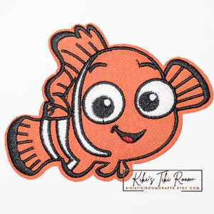 Könnte beinhalten: Gestickter orangefarbener Clownfisch-Aufnäher mit schwarzen Umrissen und weißen Streifen. Der Fisch hat große, ausdrucksstarke Augen und einen lächelnden Mund. Der Aufnäher stammt von Kiki's Tiki Room, wie durch den Text unten rechts angegeben.