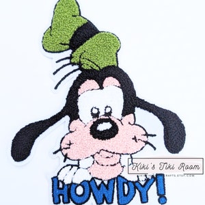 May include: Iron-on patch featuring a cartoon image of Goofy wearing a green hat and saying "Howdy!" in blue letters. The patch has a fuzzy texture.