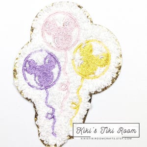 May include: Embroidered patch featuring three Mickey Mouse-shaped balloons in purple, pink, and yellow. The balloons are on a white background with a gold glitter border. The text "Kiki's Tiki Room" is at the bottom.