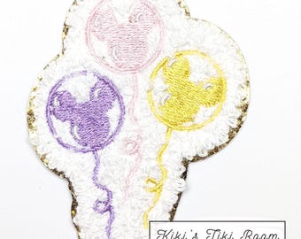 May include: Embroidered patch with three Mickey Mouse-shaped balloons in purple, pink, and yellow. The balloons are on a white background with a gold glitter border. The text "Kiki's Tiki Room" is at the bottom.