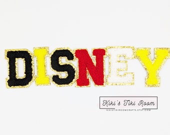 Può includere: Lettere in feltro colorate che compongono la parola "DISNEY" con bordatura glitterata dorata. Le lettere sono in vari colori: nero, giallo, rosso e bianco. L'immagine include il testo "Kiki's Tiki Room" e un indirizzo web.