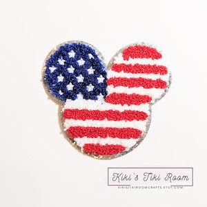 Könnte beinhalten: Eine rot-weiß-blaue amerikanische Flagge in Form eines Mickey-Mouse-Kopfes. Die Flagge besteht aus einem flauschigen, strukturierten Material und hat einen silbernen Glitzerrand.