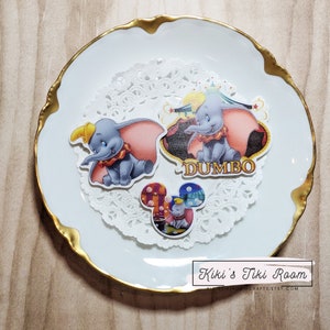 Könnte beinhalten: Drei bunte Disney-Sticker mit Dumbo dem Elefanten. Die Sticker befinden sich auf einer weißen Doily mit einem goldenen Teller dahinter. Der Text "DUMBO" befindet sich auf einem der Sticker.