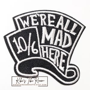 May include: A black and white embroidered patch shaped like a top hat. The patch features the white stitched text "WE'RE ALL MAD HERE" and "10/6" on a black background. The design includes a curved brim at the bottom and a tall crown structure.