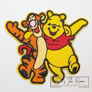 May include: Embroidered patch depicting Winnie the Pooh and Tigger embracing. Winnie the Pooh is yellow with a red top, and Tigger is orange with black stripes. The patch has a yellow border and the text "Kiki's Tiki Room" and Etsy shop URL.