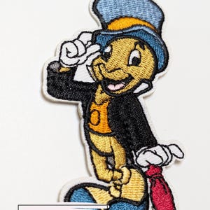 Könnte beinhalten: Gestickter Aufnäher mit einer Zeichentrick-Grille in Zylinder und Anzug, die salutiert. Die Grille ist gelb mit blauem Hut und Schuhen, einer schwarzen Jacke und einer roten Krawatte. Der Aufnäher hat unten den Text "Kiki's Tiki Room".