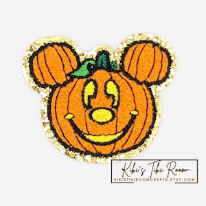 May include: A gold glitter iron-on patch featuring a smiling pumpkin with Mickey Mouse ears. The pumpkin is orange with a green stem and yellow eyes and mouth.