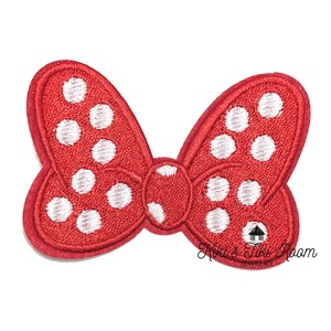 May include: Red embroidered bow with white polka dots. The bow has a rounded shape with a small center knot. The text "Kiki's Tiki Room" is printed on the lower right side of the bow.