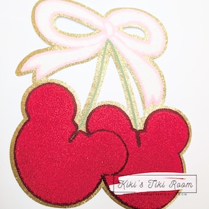 Puede incluir: Un par de cerezas rojas con contornos de purpurina dorada y un lazo rosa con detalles de purpurina dorada. Las cerezas están unidas a tallos verdes. El texto "Kiki's Tiki Room" es visible en la parte inferior de la imagen.