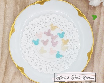 May include: A white doily with a variety of glitter-covered mouse head shapes in pastel pink, blue, and gold. The doily is on a white plate with a gold rim.