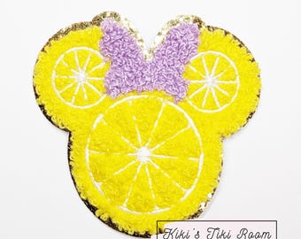 Könnte beinhalten: Gelbe, gestickte Zitronenscheiben aus Filz in Form von Mickey Mouse Ohren mit einer lila Schleife in der Mitte. Die Applikation hat einen goldenen Glitzer-Umrandung.