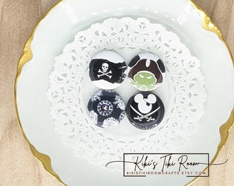 May include: Four round buttons with different designs. The buttons are on a white doily on a white plate with a gold rim. The designs include a skull and crossbones, a cartoon pirate, a floral design, and Mickey Mouse with a pirate sword. The text "Kiki's Tiki Room" and "kikistikikroomcrafts.etsy.com" is printed on the plate.