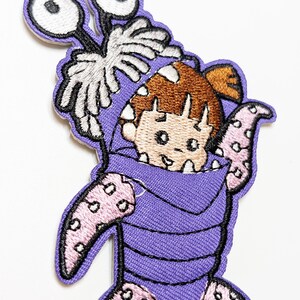 May include: Embroidered patch featuring a cartoon character in a purple monster costume. The patch has a light purple background with a detailed design of the character's face, arms, and legs. The character has brown hair and large white eyes.