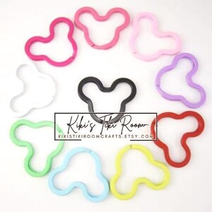 May include: A collection of colorful, Mickey Mouse-shaped keychains in various colors, including pink, red, green, blue, yellow, purple, white, and black. The keychains are arranged in a circular pattern. The text "Kiki's Tiki Room" is visible.