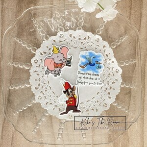May include: A clear, scalloped-edge glass plate holds three cartoon character stickers on a white doily. The stickers feature Dumbo the elephant, a stork carrying a baby, and a mouse in a red uniform. Text on the stork sticker reads, "Straight from heaven up above, here is a baby for you to love."