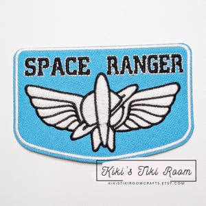 Może przedstawiać: Jasnoniebieska naszywka z haftem z napisem "SPACE RANGER" czarnymi literami. Naszywka przedstawia biało-czarny stylizowany emblemat rakiety ze skrzydłami. Naszywka ma białą obwódkę.