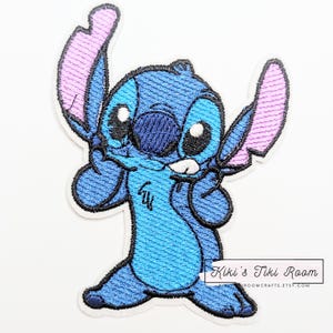 Pode incluir: Patch bordado com o personagem Stitch de Lilo & Stitch. O patch é principalmente azul com orelhas internas rosa e contornos pretos. O personagem está em uma pose brincalhona. O patch tem uma borda branca e o texto "Kiki's Tiki Room" é visível.