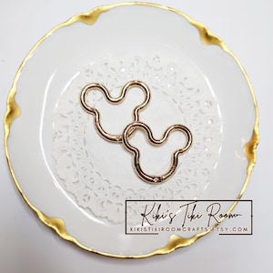 Puede incluir: Dos cierres metálicos dorados con forma de Mickey Mouse descansan sobre un mantelito blanco sobre un plato blanco con borde dorado. Los cierres están abiertos y listos para usar. El texto "Kiki's Tiki Room" es visible.