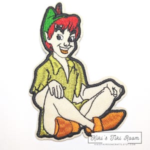 May include: Embroidered patch of Peter Pan in a seated position. The patch depicts Peter Pan with red hair, a green hat, and a yellow shirt. The patch is outlined in black and includes the text "Kiki's Tiki Room" at the bottom.
