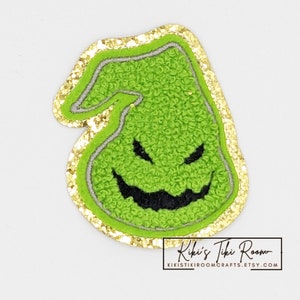 Op de afbeelding: Groen vilt karakter patch met een zwarte gestikte glimlach en ogen. De patch is omgeven door een gouden glitterrand.