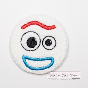 Könnte beinhalten: Weißer Plüsch-Patch mit einem gestickten Comic-Gesicht. Das Gesicht hat rote Augenbrauen, schwarze Augen und ein blaues Lächeln.