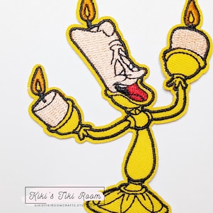 Può includere: Toppa ricamata raffigurante un personaggio di candelabro giallo con un volto sorridente, lingua rossa e tre candele accese. La toppa ha un contorno nero e il testo "Kiki's Tiki Room" in basso. Lo sfondo è bianco.