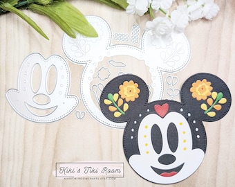 May include: A die-cut template for crafting a Mickey Mouse head with a Day of the Dead theme. The template features a black and white Mickey Mouse head with floral details and a sugar skull design. The template is shown on a wooden surface with a white floral background.