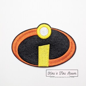 Könnte beinhalten: Gestickter Aufnäher mit dem Logo des Animationsfilms Die Unglaublichen. Der Aufnäher hat eine schwarze ovale Form mit einem orangefarbenen Rand, einem gelben Rechteck und einem gelben Kreis mit weißem Zentrum. Der Aufnäher ist von Kiki's Tiki Room.
