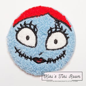 May include: A blue and red embroidered patch featuring Sally from the Nightmare Before Christmas. The patch is made of a soft, fuzzy fabric and has a stitched smile and black eyes.