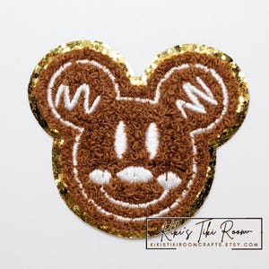May include: A brown embroidered patch in the shape of a cartoon mouse head with white details and a gold sequin border. The patch has a smiling face and the text "Kiki's Tiki Room" at the bottom.
