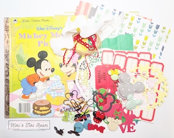 May include: A collection of Disney-themed scrapbook supplies. Includes a "Mickey Mouse Picnic" Little Golden Book, red tickets, paper cutouts, and decorative elements. The color palette features red, yellow, and pastel colors.