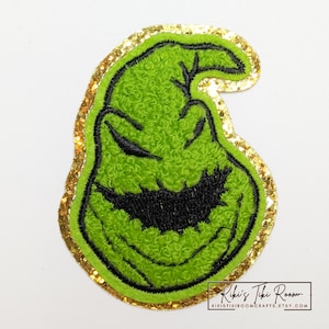 May include: A green, textured patch depicting a cartoon face with a black smile and eyes. The patch is outlined in black and has a gold glitter border. The text "Kiki's Tiki Room" is visible in the bottom right corner.